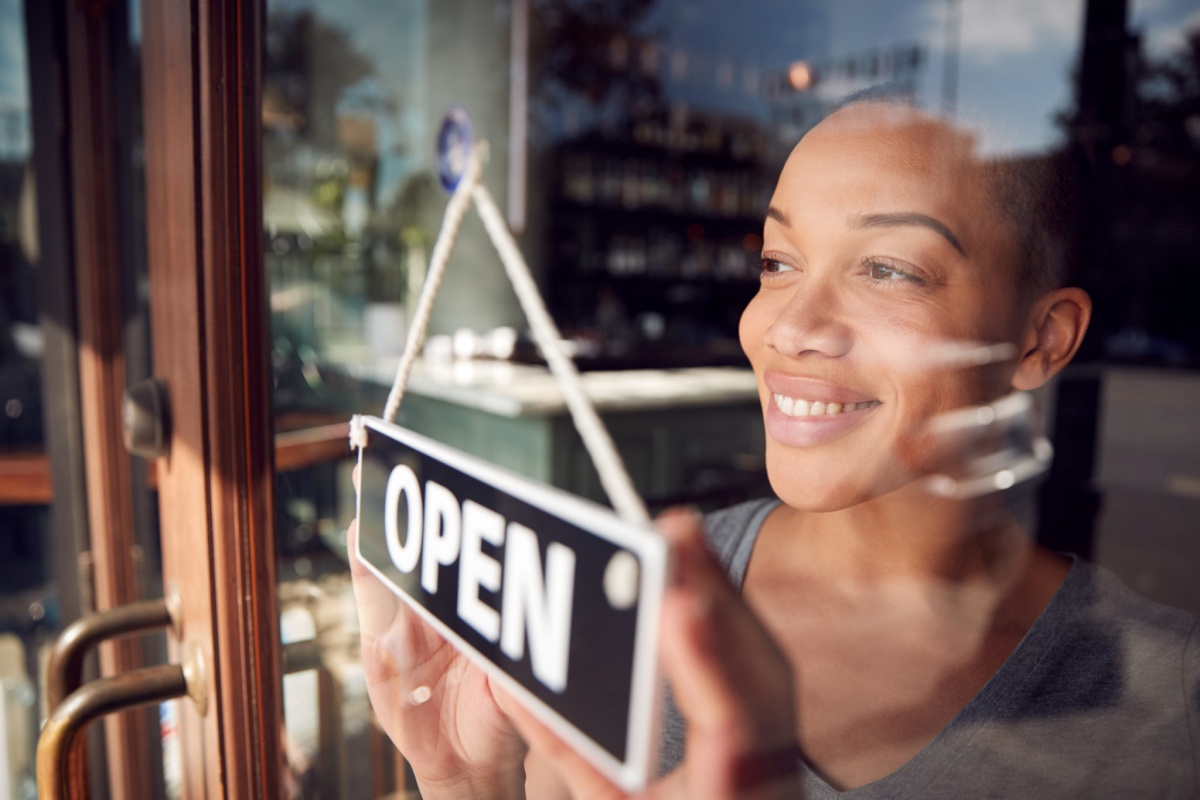 Black woman turning the "open" sign on a glass business door, representing changing trends in the restaurant business